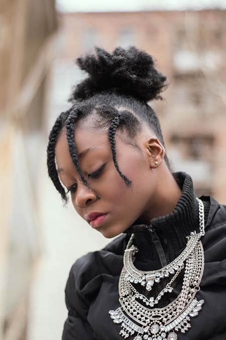 Black womxn with a professional afro-textured puff with 4-5 box braids as a fringe hairstyle wearing a big silver necklace