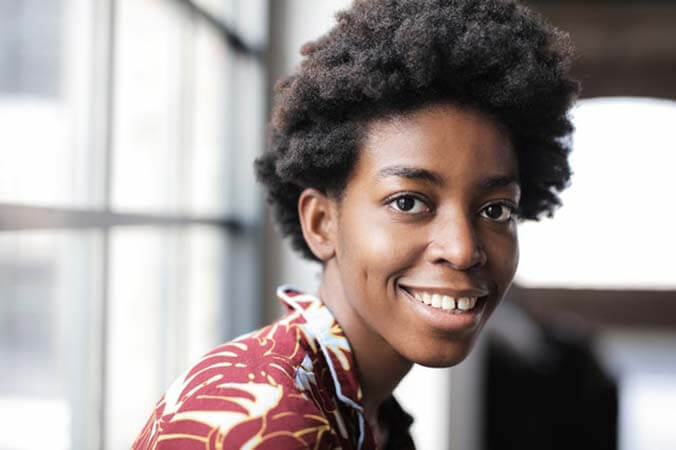 Black womxn with a professional short-mid length 4C afro hairstyle sitting by the window, wearing a floral shirt and smiling