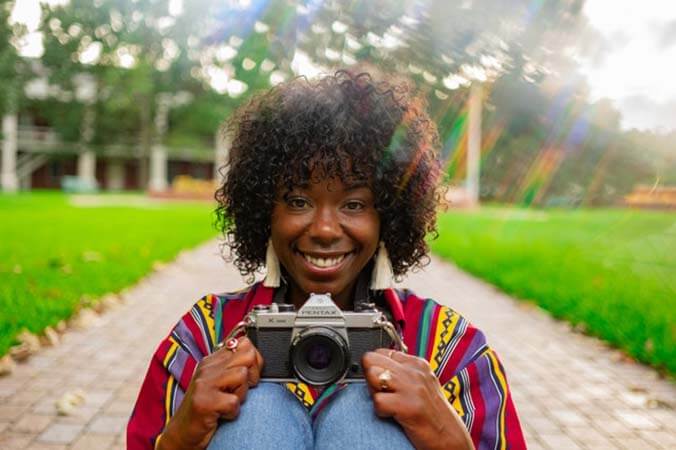 Black womxn with a professional curly afro hairstyle, sitting, holding a camera on her knees, and smiling