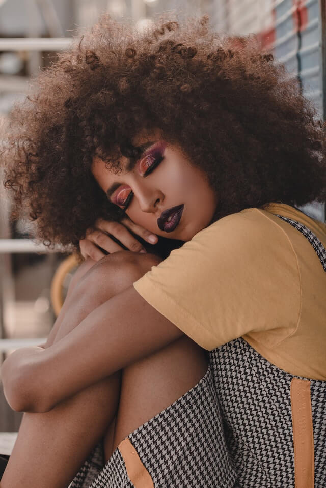 Black womxn with professional big afro, dark purple lipstick, smokey eyes, resting her head on her knees with closed eyes
