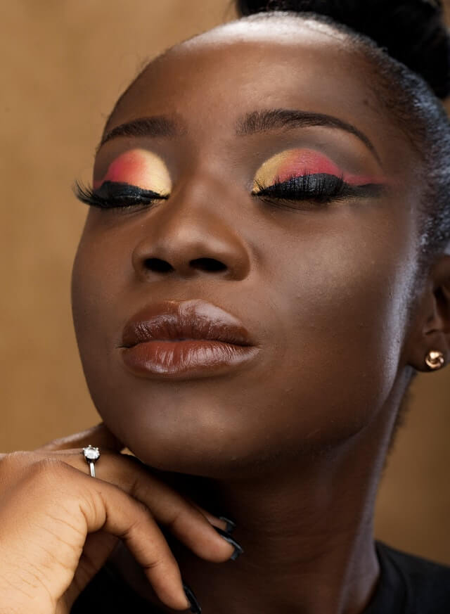 Black womxn with workplace makeup, black eyeliner, yellow and orange eyeshadow, rust red lips, head raised with closed eyes