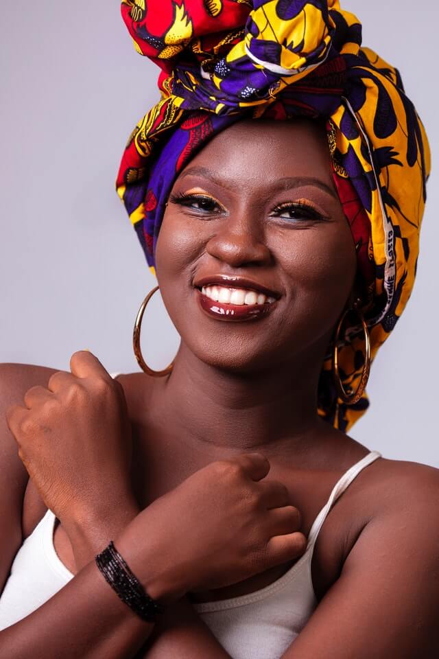 Black womxn with turban, workplace makeup, dark red lipgloss and orange eyeshadow, laughing and looking to the camera