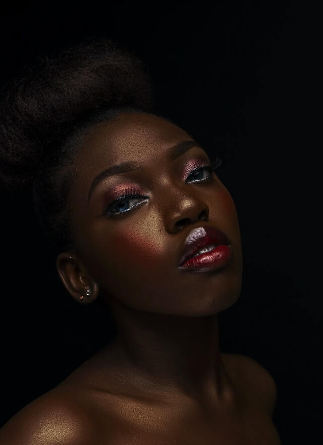 Black womxn with workplace makeup, dark red lipgloss, orange blush and eyeshadow, holding her head up, looking to the camera
